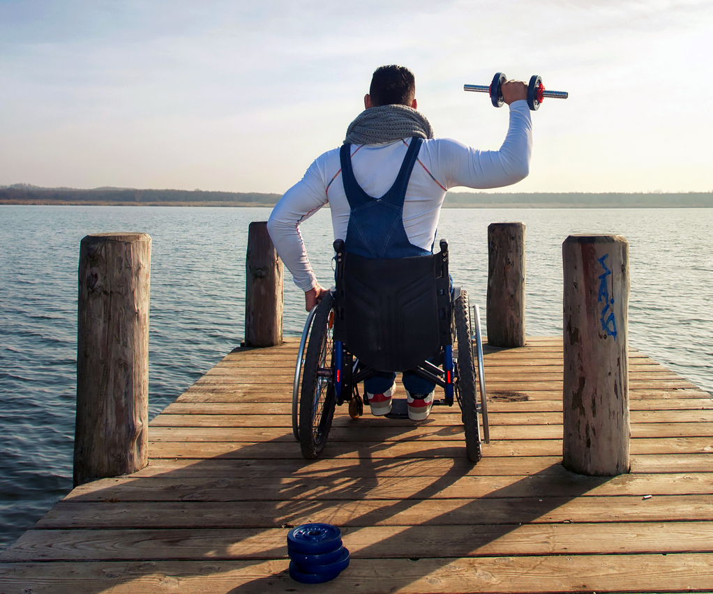 Young male wheelchair user on a pier exercising with a weight in his right hand.